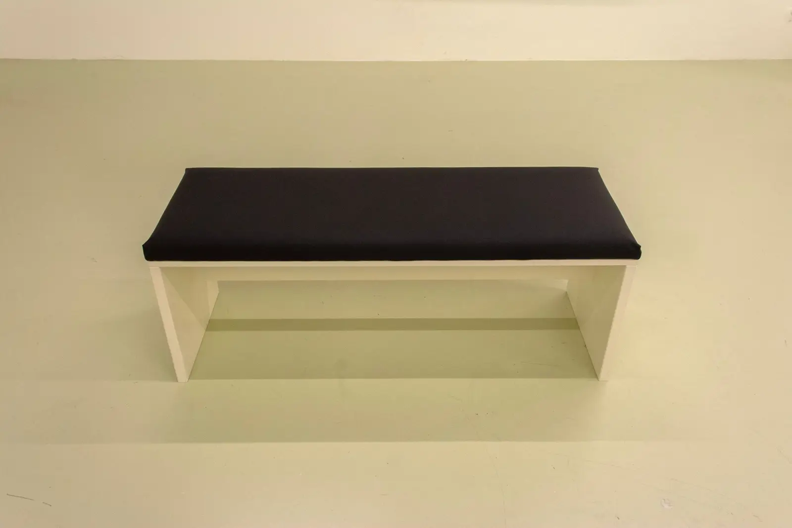A plain wooden gallery bench with a dark cushion against a pale wall